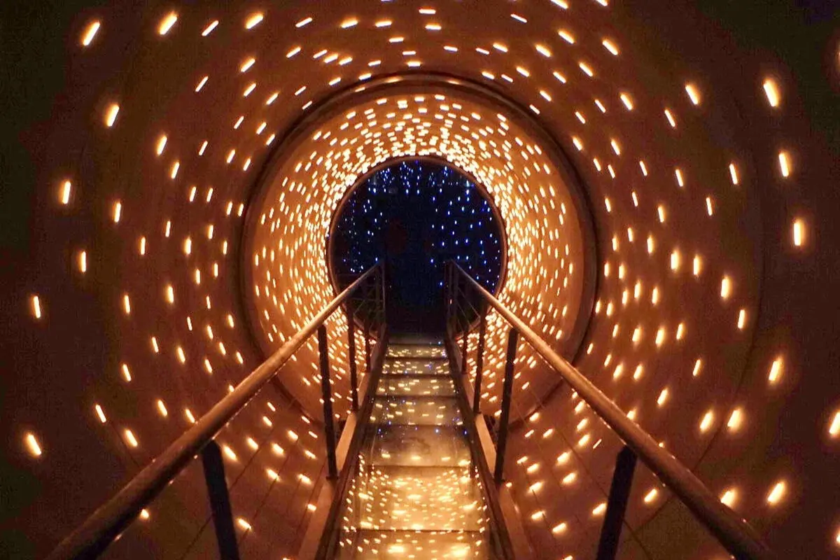 Une passerelle avec des rampes à l'intérieur d'un tunnel éclairé par de nombreuses petites lumières chaudes, créant un effet étoilé.