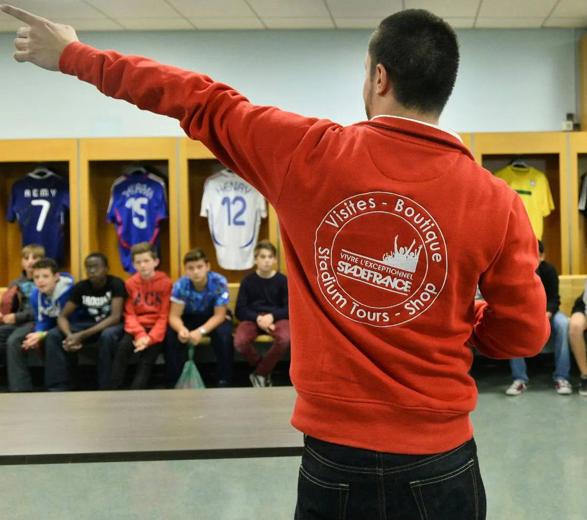Un adulte vêtu d'une veste rouge fait visiter un groupe de jeunes assis dans un vestiaire, les maillots suspendus derrière eux.