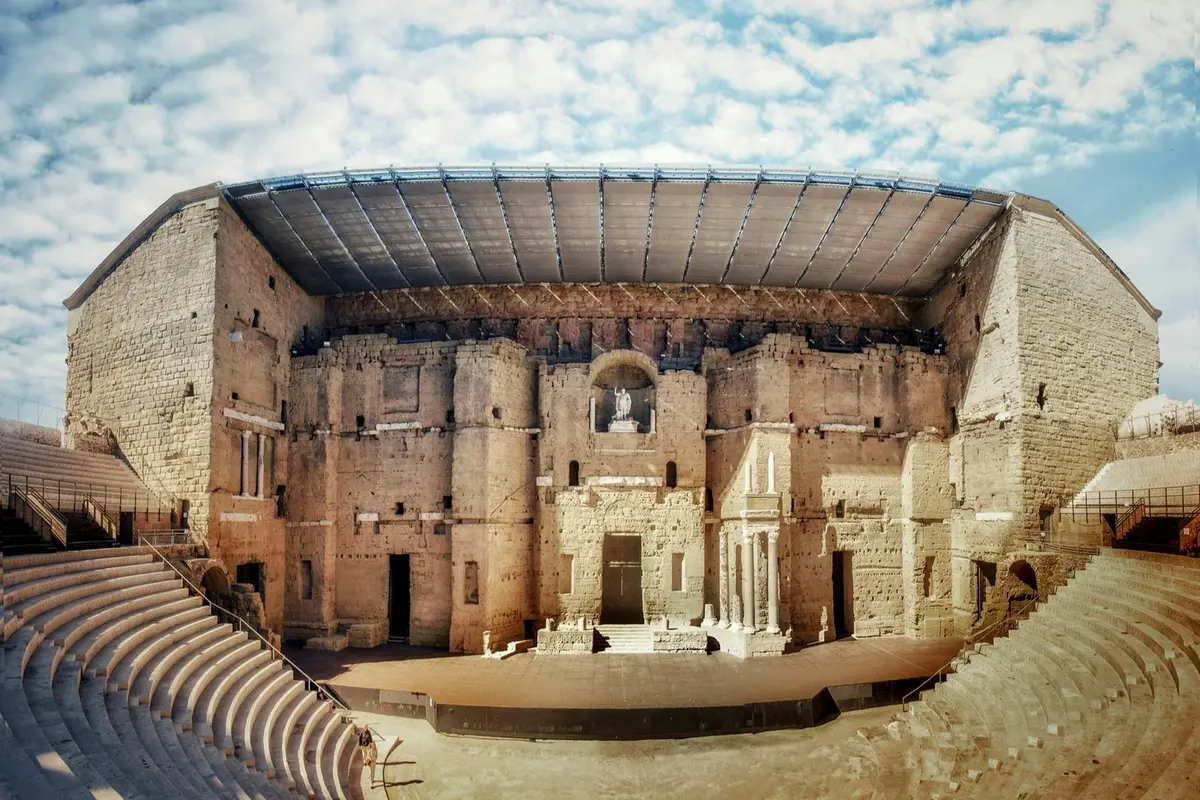Un amphithéâtre antique en pierre, doté d'une façade richement décorée, d'une scène et de gradins, sous un ciel partiellement nuageux.