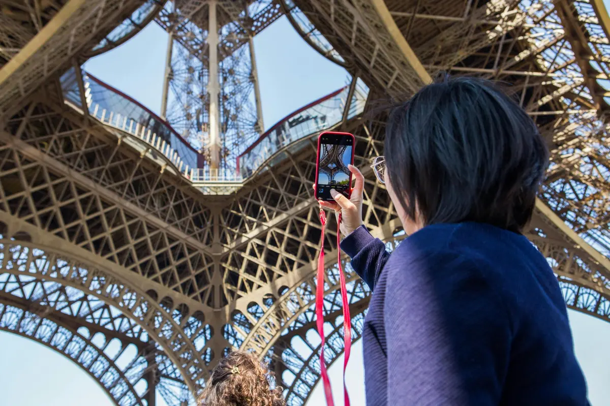A person holding a smartphone taking a photo of the intricate underside of the Eiffel Tower on a clear day.