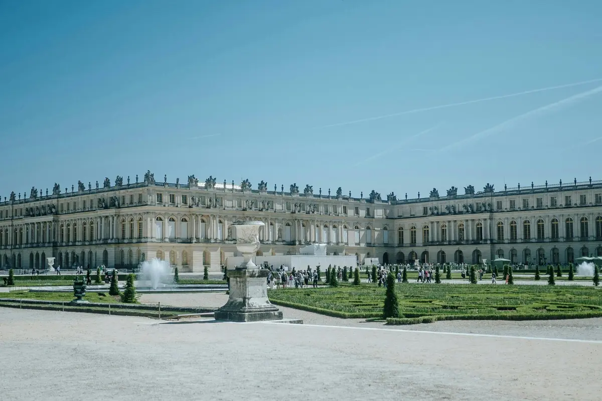 Un grand palais à l'architecture ornée, un jardin formel avec des fontaines au premier plan, et des visiteurs qui se promènent.