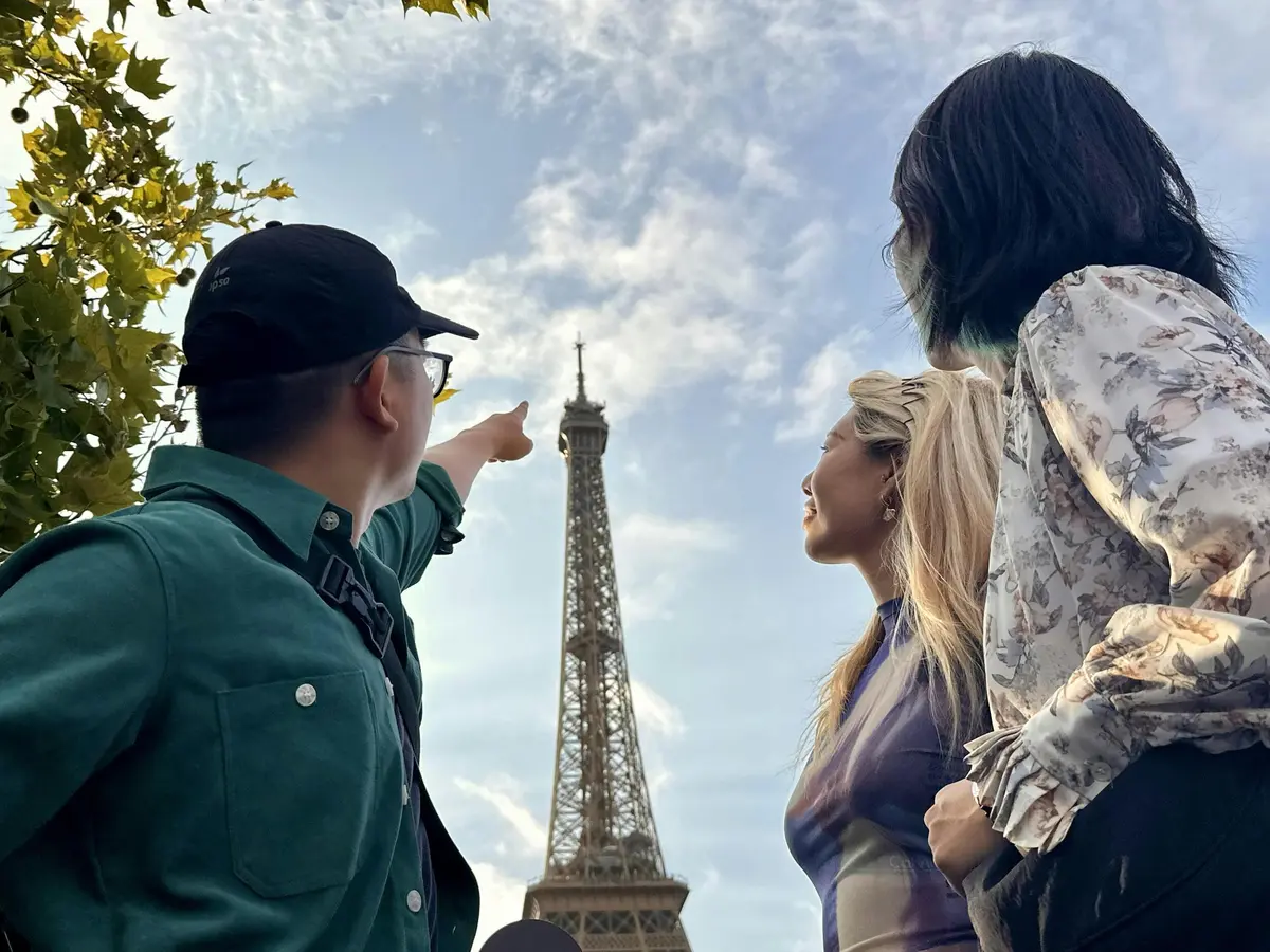Trois personnes se tiennent près de la Tour Eiffel. L'une d'entre elles pointe le doigt vers le haut, créant ainsi un sentiment de crainte. Le ciel est partiellement nuageux.