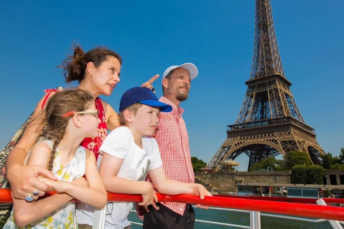 Deux adultes et deux enfants sur un bateau, avec la Tour Eiffel en arrière-plan dans un ciel bleu clair.