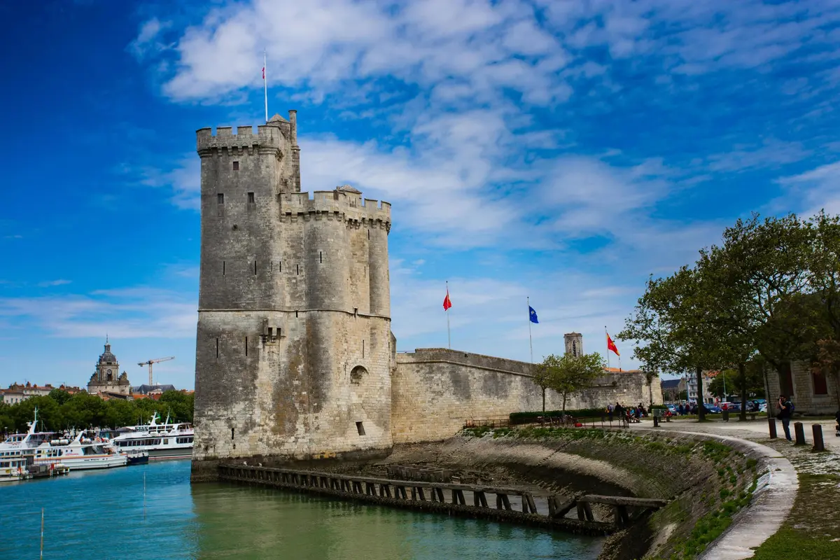 Forteresse historique en pierre avec drapeaux et tours crénelées au bord d'une eau calme et verdâtre sous un ciel partiellement nuageux.
