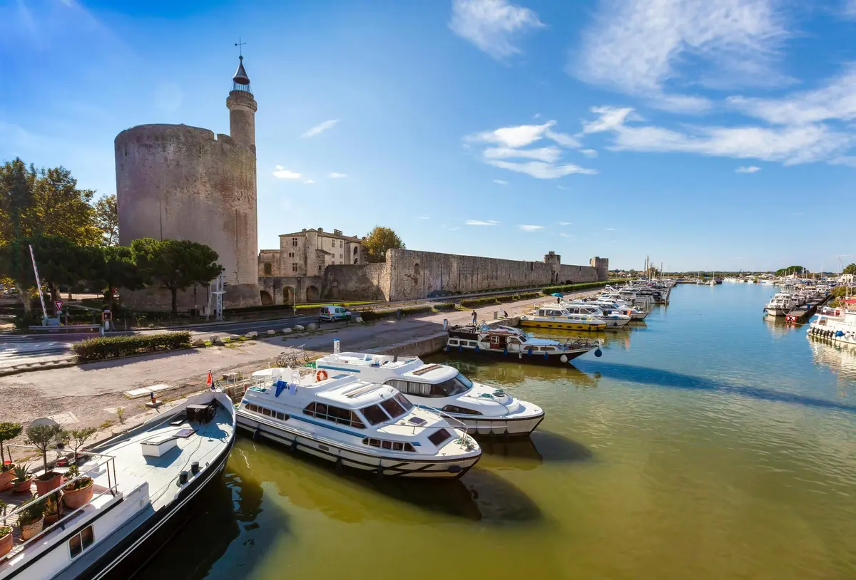 Bateaux amarrés le long d'un front de mer avec un château en pierre et un mur de forteresse en arrière-plan sous un ciel bleu clair.