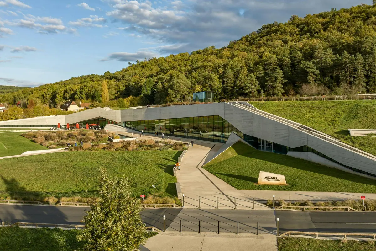Un bâtiment de musée moderne avec un toit vert et des murs en verre, entouré de verdure et de sentiers, sous un ciel partiellement nuageux.
