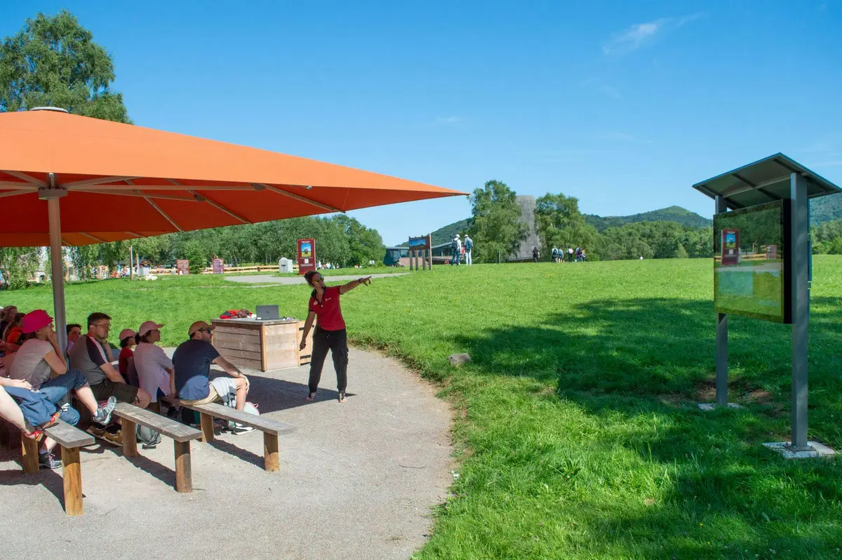L'instructeur pointe du doigt tout en s'adressant à un groupe assis sous un auvent orange dans un espace extérieur herbeux par une journée ensoleillée.