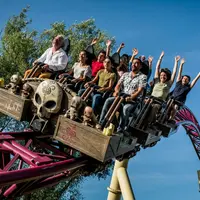 Les passagers d'une montagne russe lèvent les mains et crient d'excitation. Le manège est orné de crânes et de motifs complexes. Le ciel est d'un bleu limpide. &copy; Walibi Rhone Alpes