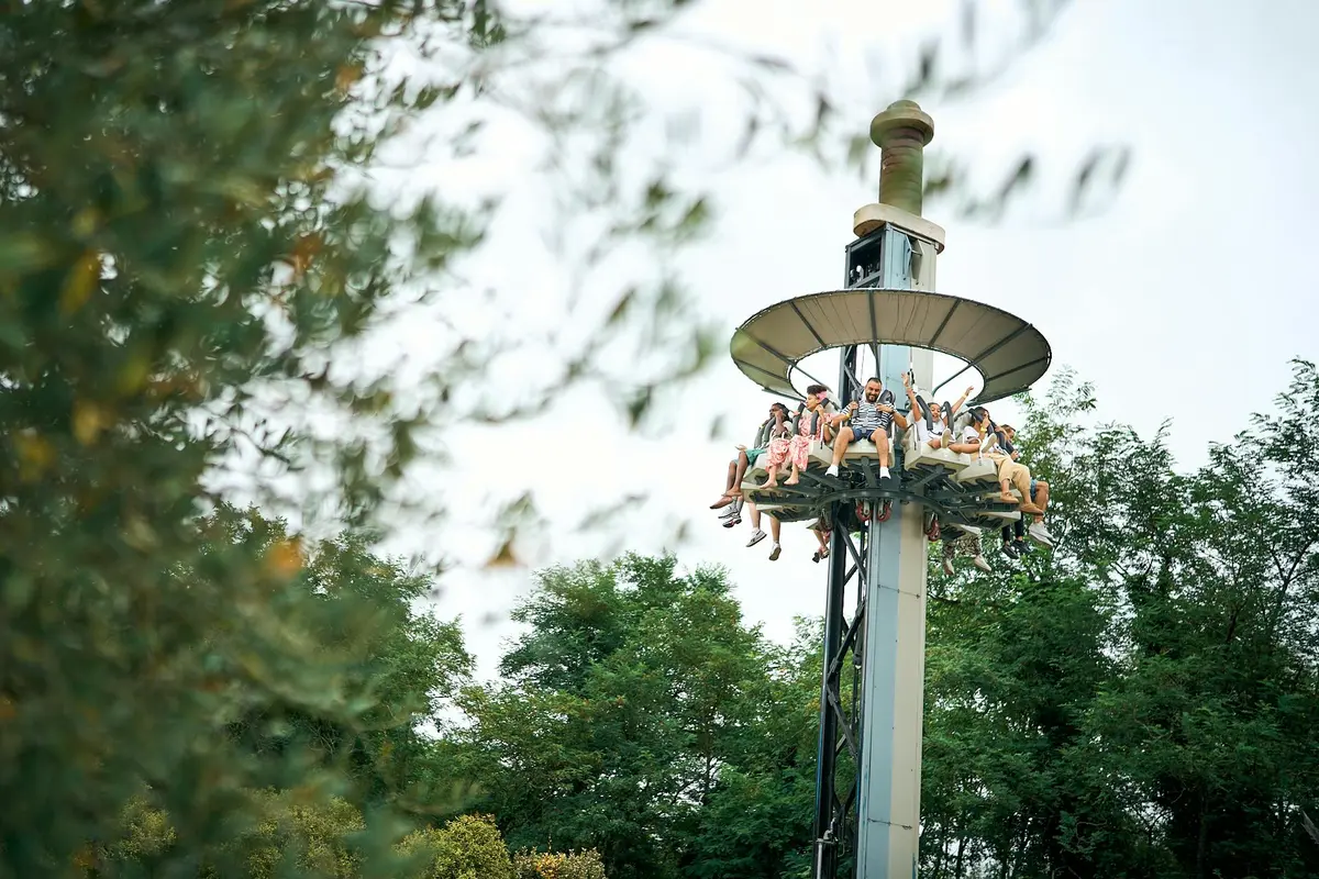 Personnes assises sur un manège surélevé dans un parc d'attractions, entouré de verdure.