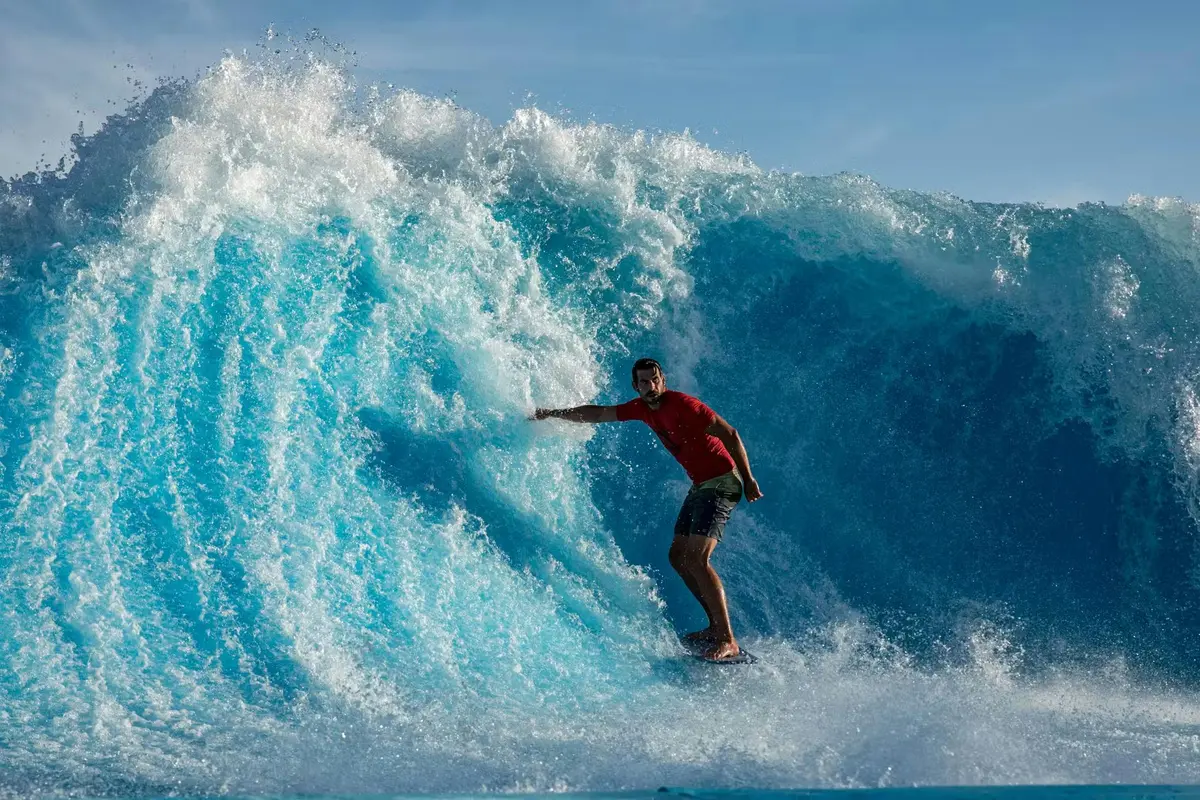Un surfeur chevauchant une grande vague bleue sous un ciel dégagé, vêtu d'un t-shirt rouge et d'un short.