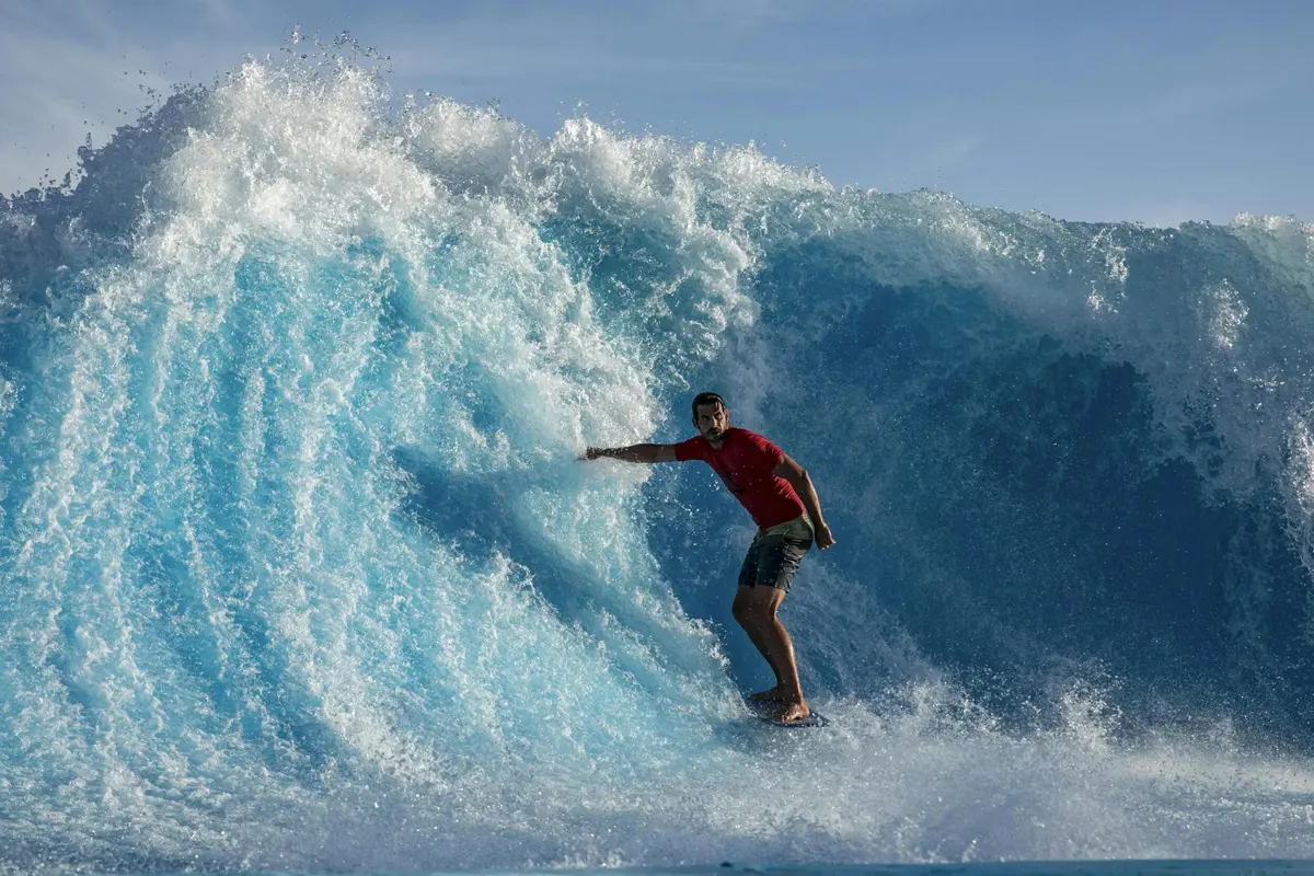 Personne vêtue d'une chemise rouge et d'un short qui surfe sur une grande vague bleue de l'océan sous un ciel dégagé.