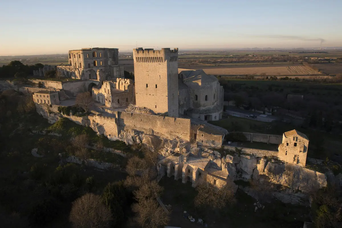 Vue aérienne d'un château médiéval sur une colline avec des champs et des montagnes lointaines sous un ciel clair.