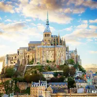 Une abbaye historique en pierre avec une haute flèche située sur une colline, entourée de bâtiments plus petits sous un ciel partiellement nuageux. DR