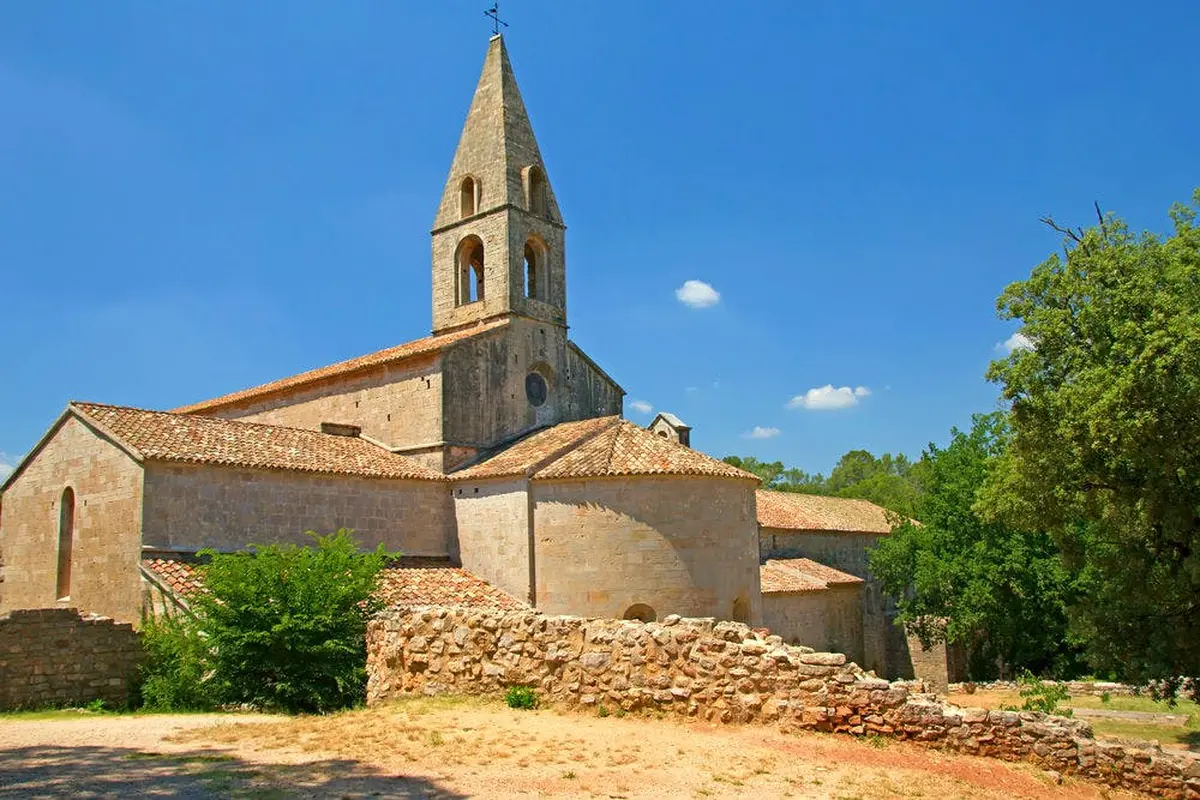 Église en pierre avec un haut clocher pointu et un toit en tuiles, entourée de verdure et d'un muret de pierre sous un ciel bleu.