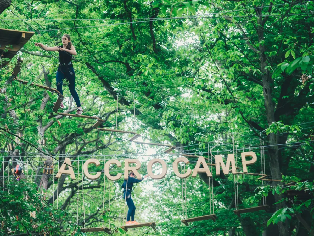 Deux personnes naviguent sur un parcours de cordes dans une forêt luxuriante, avec de grandes lettres épelant ACCROCAM suspendues au premier plan.