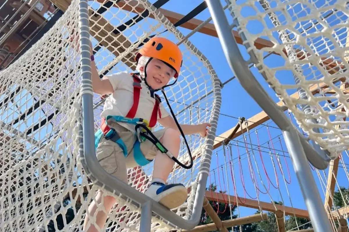 Un enfant portant un casque et un harnais escalade un filet de parcours de corde par une journée ensoleillée.