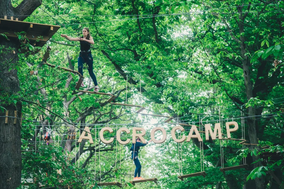 Deux personnes évoluent dans un parcours de cordes au milieu d'arbres verdoyants ; l'une se tient sur une planche, tandis que l'autre se tient en équilibre près de lettres en bois épelant ACCRO.