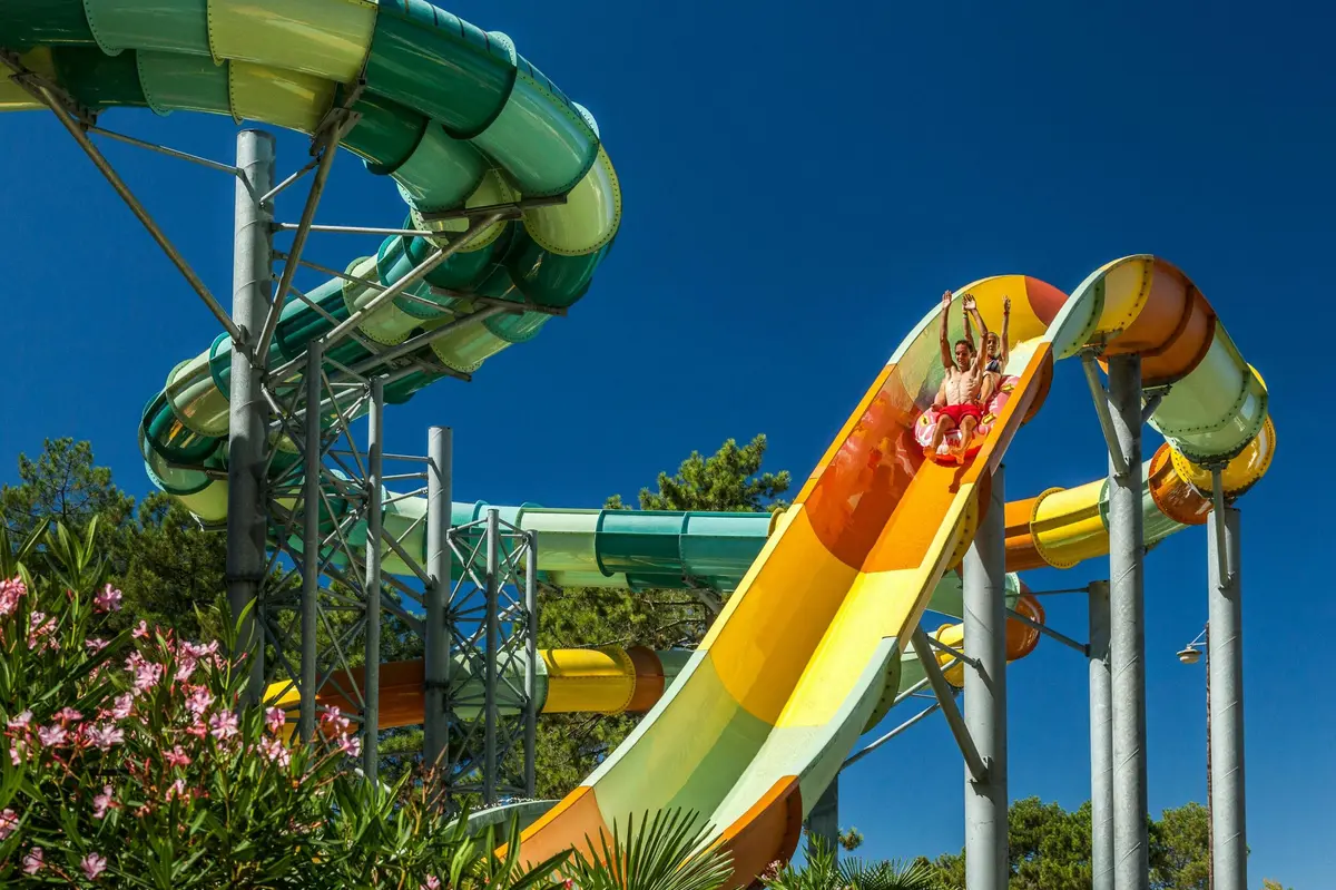 Deux personnes d&eacute;valant un toboggan aquatique color&eacute; dans un parc aquatique, sous un ciel bleu limpide, au milieu d'un cadre verdoyant.