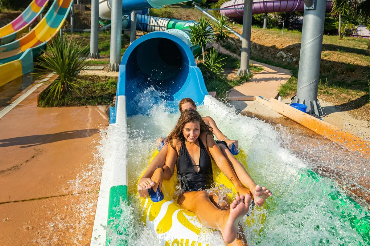 Deux personnes descendent un toboggan aquatique sur un radeau gonflable jaune et s'éclaboussent en sortant du toboggan à l'extérieur.