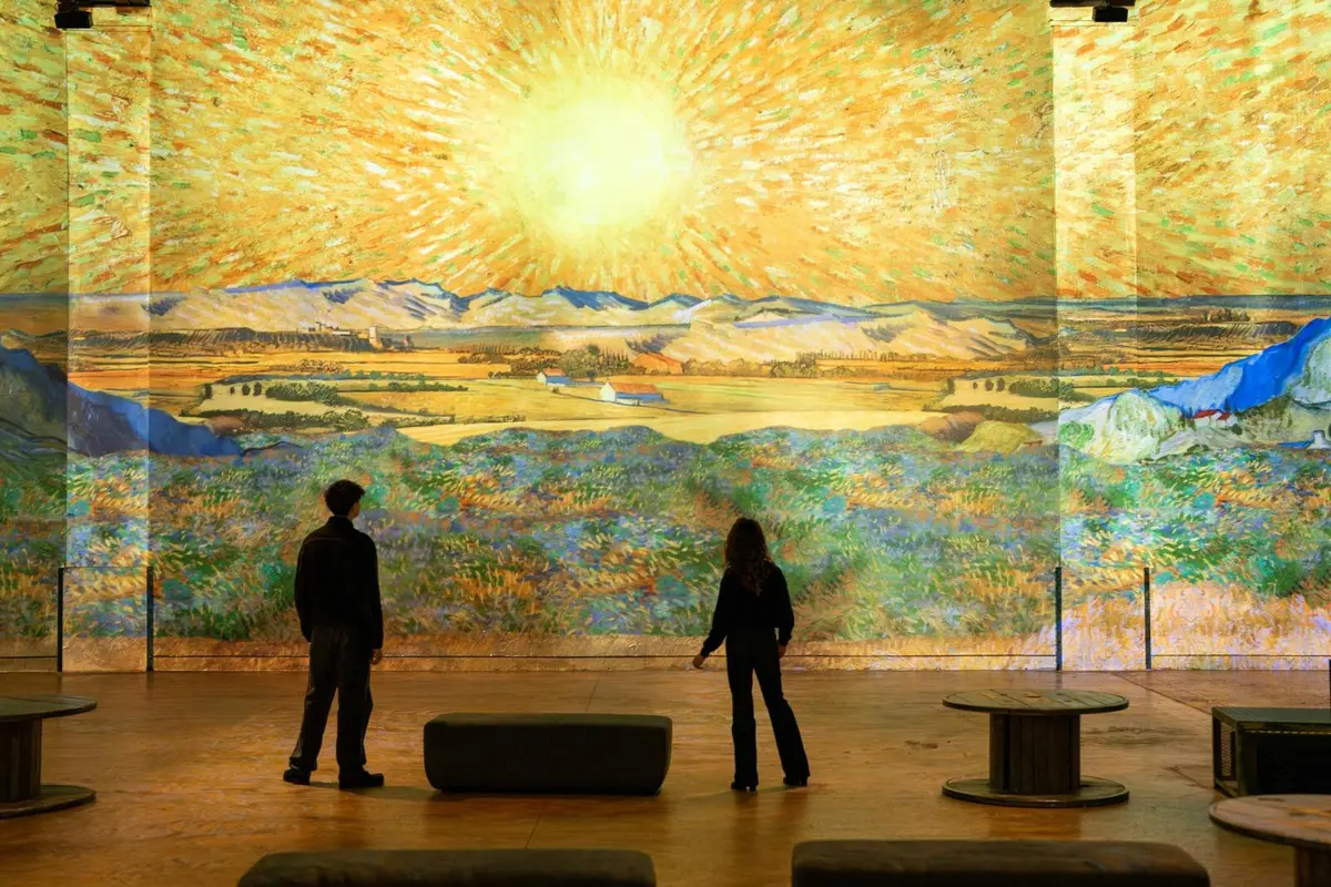 Deux personnes admirent une grande œuvre d'art lumineuse représentant un paysage champêtre plein de vie, baigné par un soleil éclatant.
