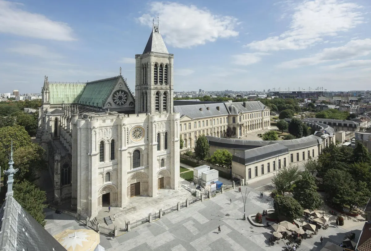Vue aérienne d'une grande cathédrale historique avec une haute tour d'horloge à côté d'une vaste cour et de bâtiments adjacents sous un ciel partiellement nuageux.