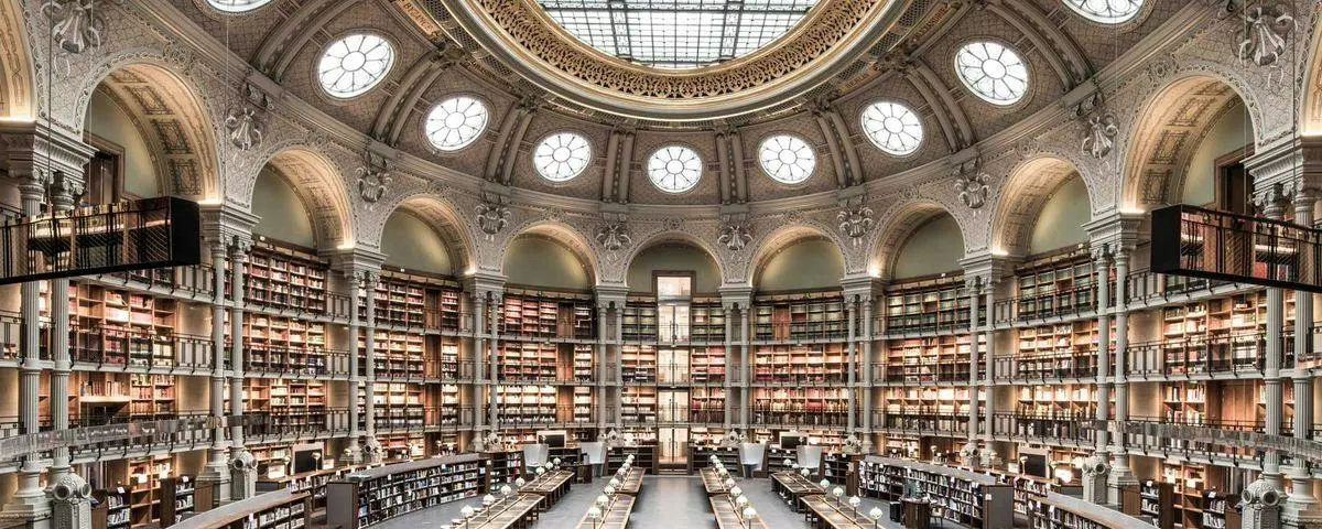 L'intérieur d'une grande bibliothèque, avec un plafond en dôme, des détails richement décorés et des étagères remplies de livres. De grands bureaux se trouvent en contrebas.