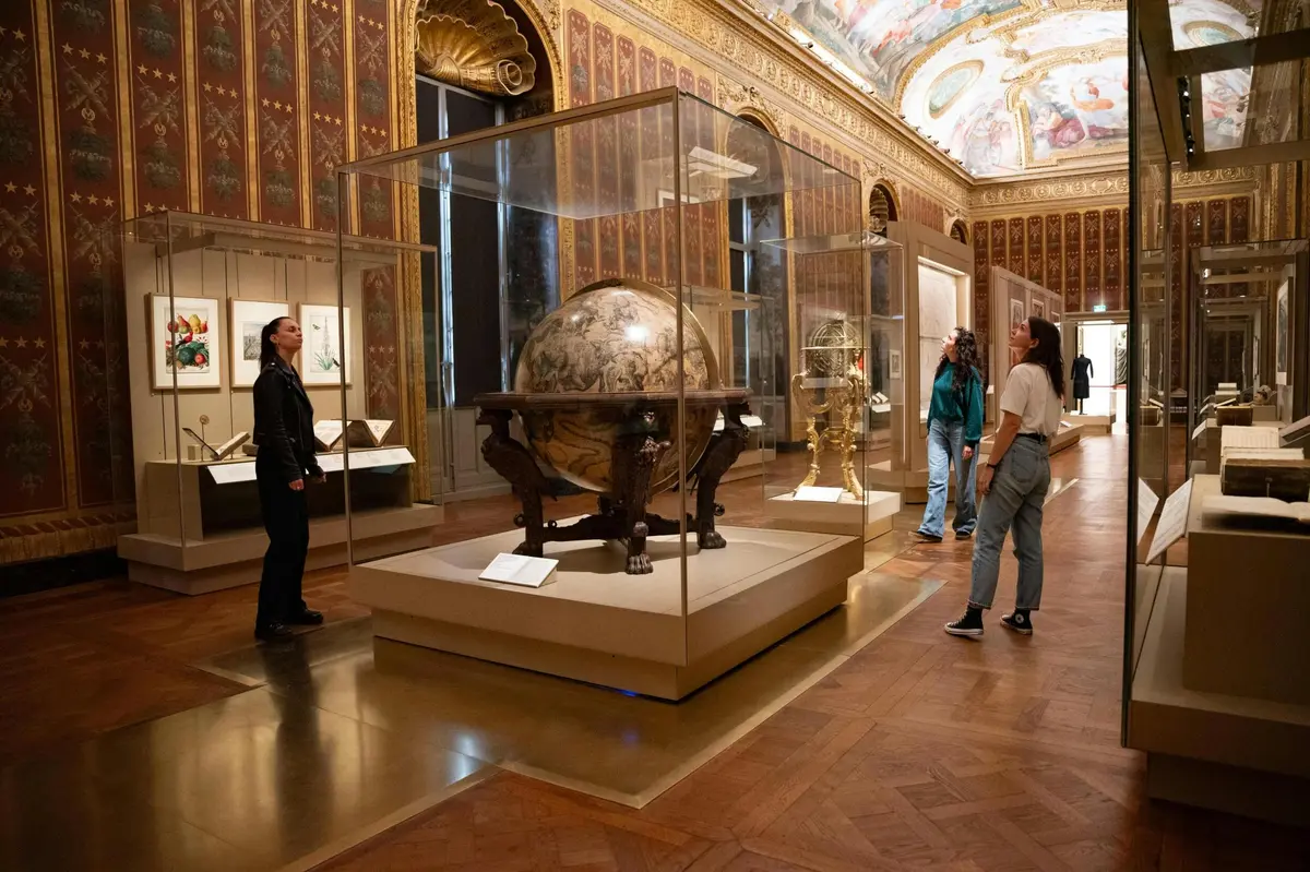 Four people admire artifacts, including a large globe, in a museum with ornate walls and a decorative ceiling.