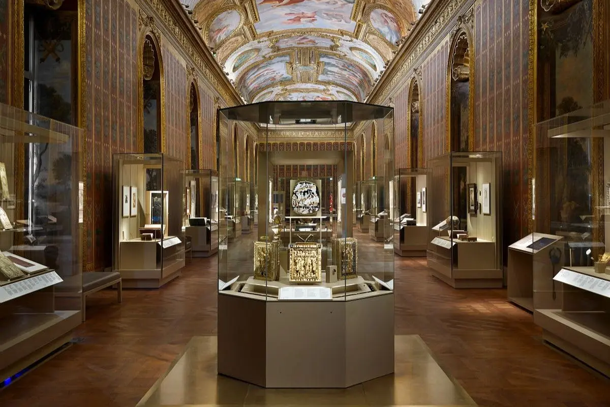 Une galerie de musée avec des vitrines ornées, des œuvres d'art sur les murs et un plafond peint avec des motifs complexes.