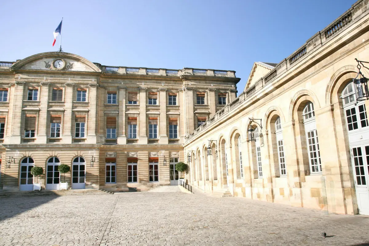 A large, historic building with arched windows and a paved courtyard under a clear blue sky.