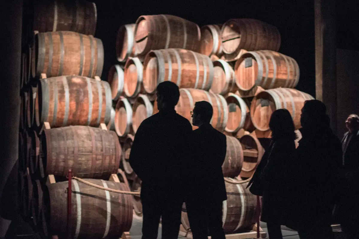 Trois personnes observent des piles de grands tonneaux en bois dans un endroit faiblement éclairé.