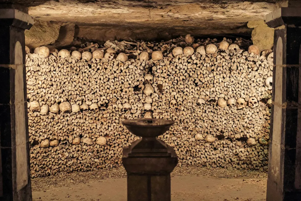 Une pièce dont les murs sont constitués d'os et de crânes humains empilés, avec, au premier plan, un piédestal sur lequel repose un bol.
