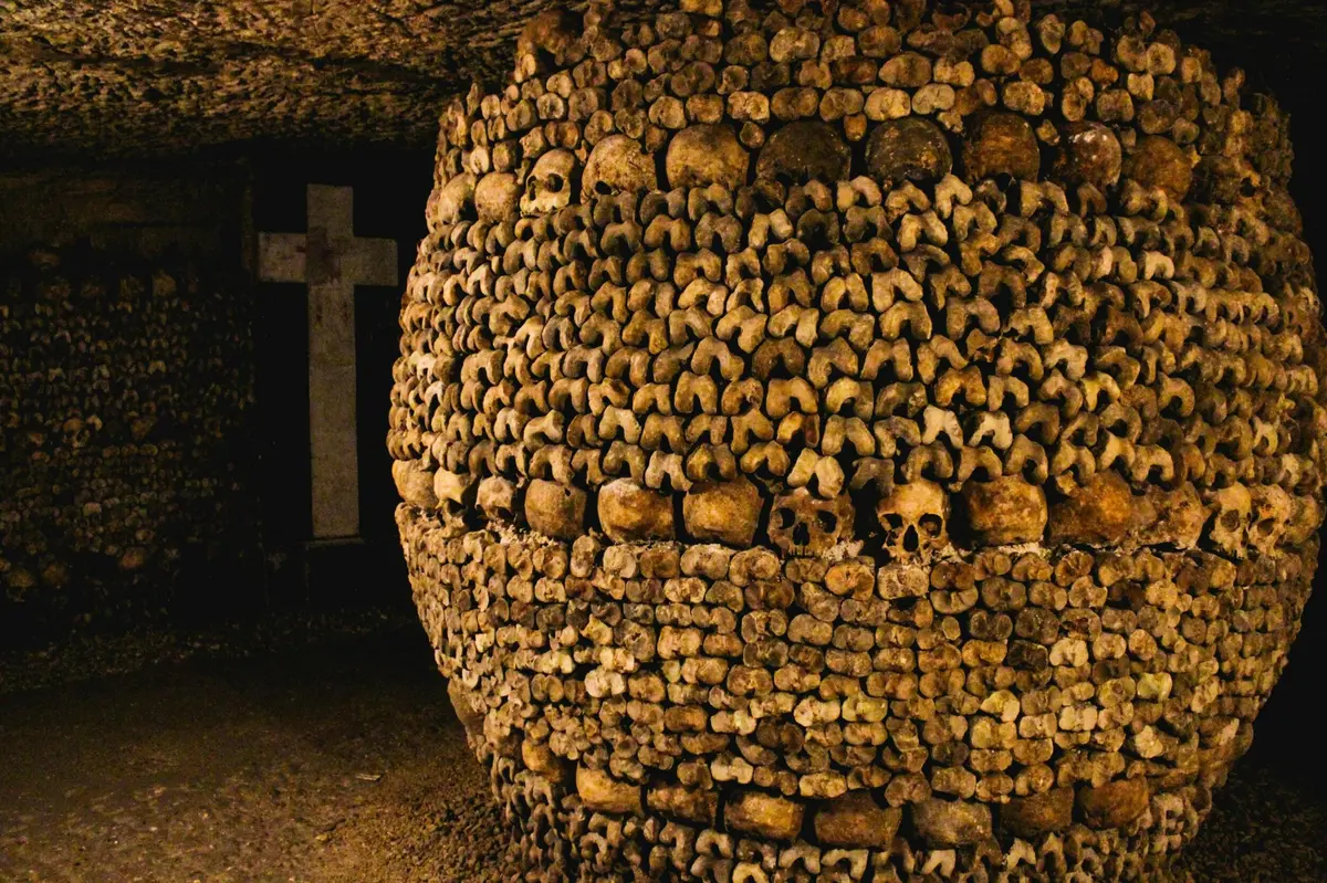 Mur composé d'ossements et de crânes humains disposés dans un ossuaire souterrain, avec une croix visible à l'arrière-plan.