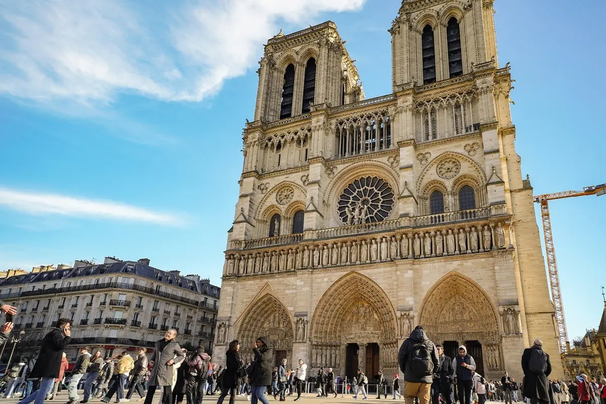 Personnes marchant devant la cathédrale Notre-Dame de Paris par temps clair, avec quelques bâtiments visibles sur la gauche.