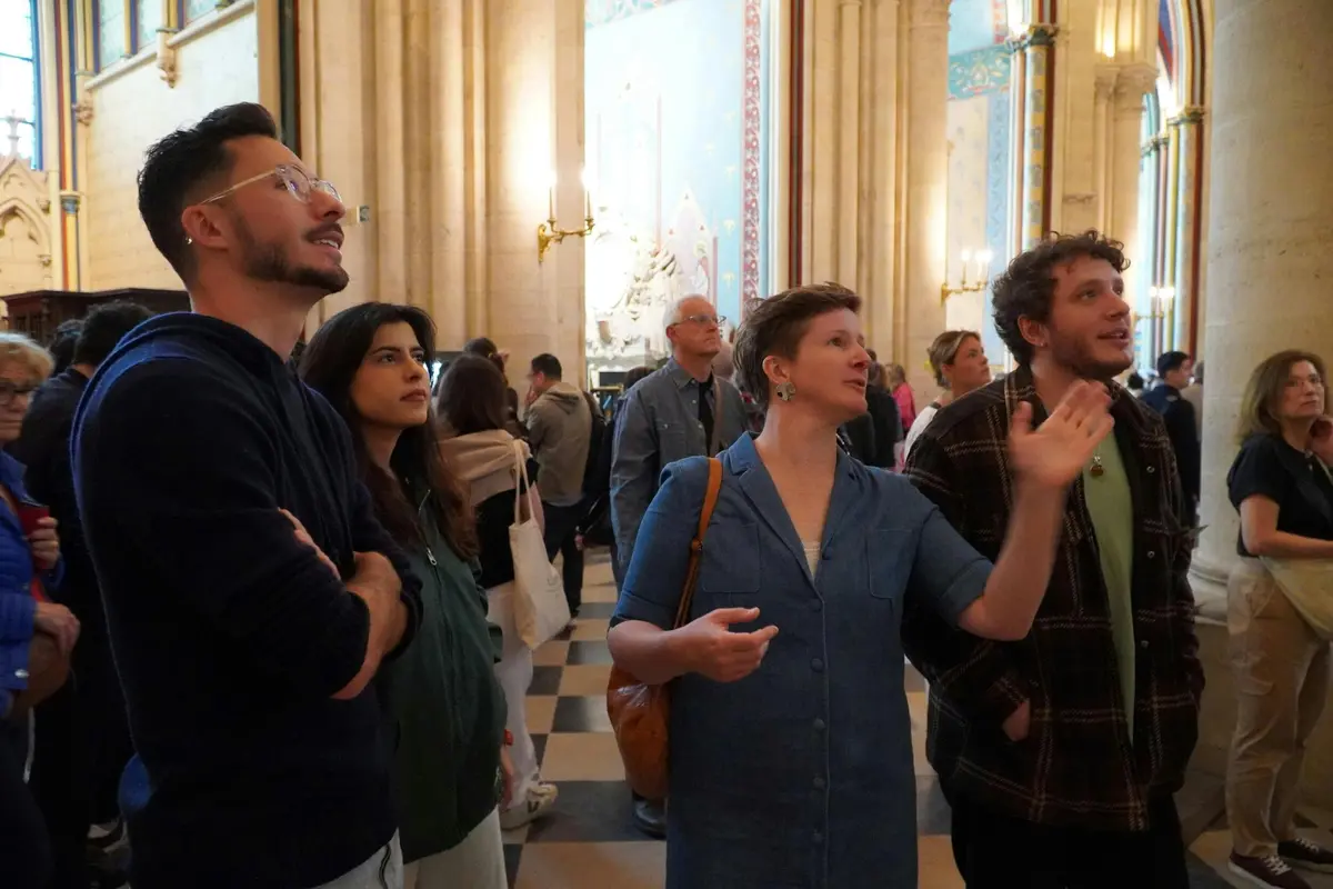 Des personnes observent des œuvres d'art dans un cadre semblable à une cathédrale, avec de hauts plafonds et un décor orné. Une personne pointe du doigt tandis que d'autres regardent.