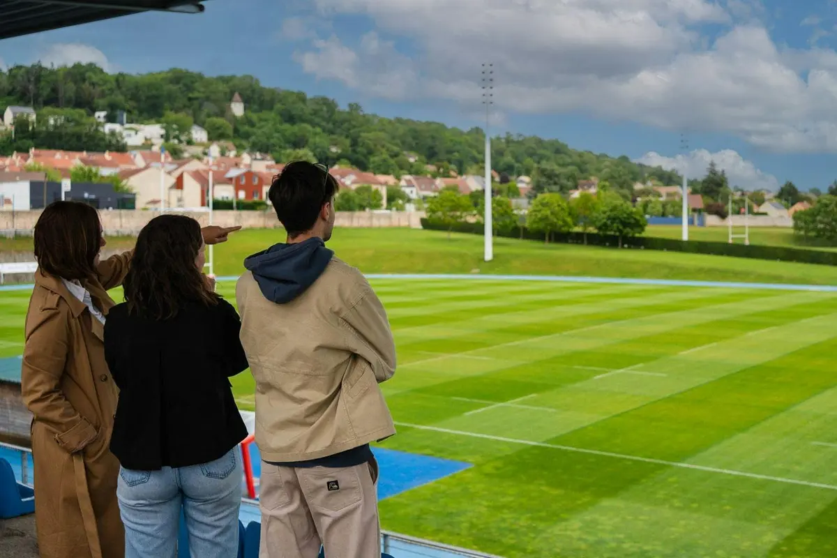 Deux personnes se tenant sur une terrasse donnant sur un terrain de sport, avec en arrière-plan un village pittoresque et des arbres.