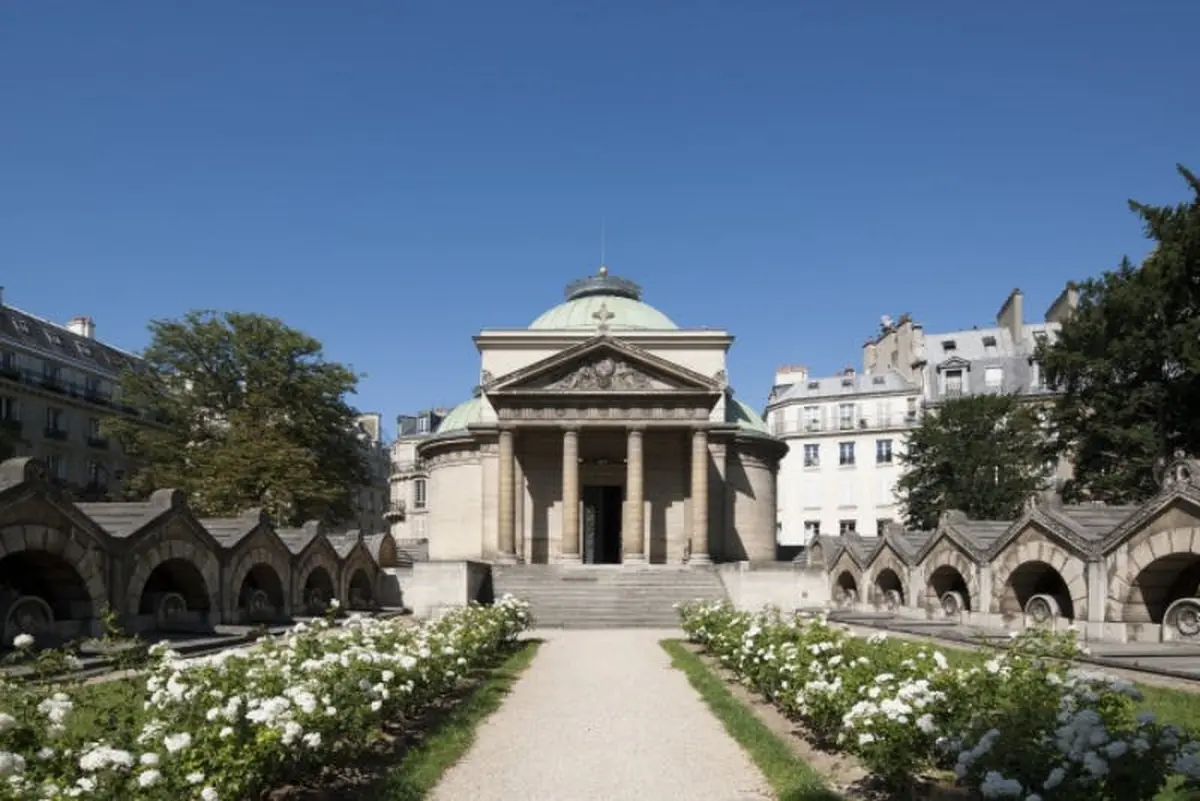 Un bâtiment historique avec un dôme et des colonnes se dresse au milieu d'un jardin bien entretenu avec des fleurs blanches et un chemin de gravier sous un ciel clair.
