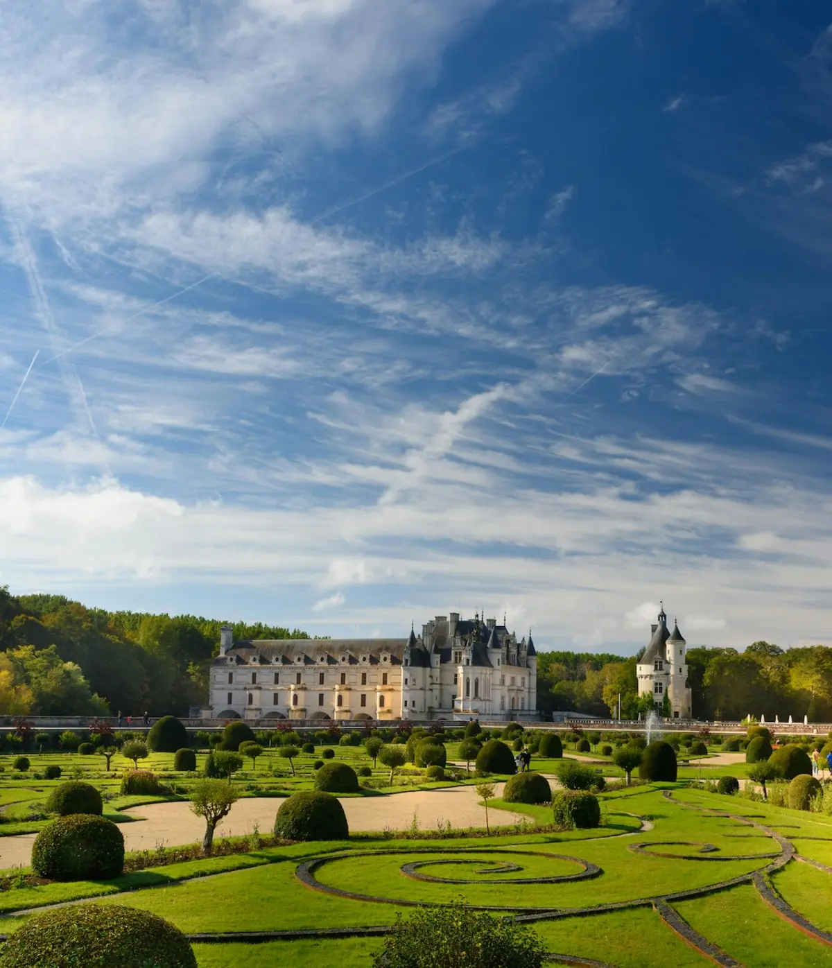 Un ch&acirc;teau pittoresque avec des jardins paysagers sous un ciel bleu avec des nuages &eacute;pars et des arbres en arri&egrave;re-plan.