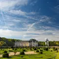 Un château pittoresque avec des jardins paysagers sous un ciel bleu avec des nuages épars et des arbres en arrière-plan.