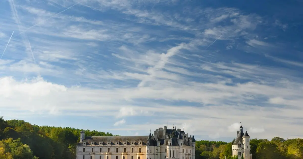 Un château pittoresque avec des jardins paysagers sous un ciel bleu avec des nuages épars et des arbres en arrière-plan.
