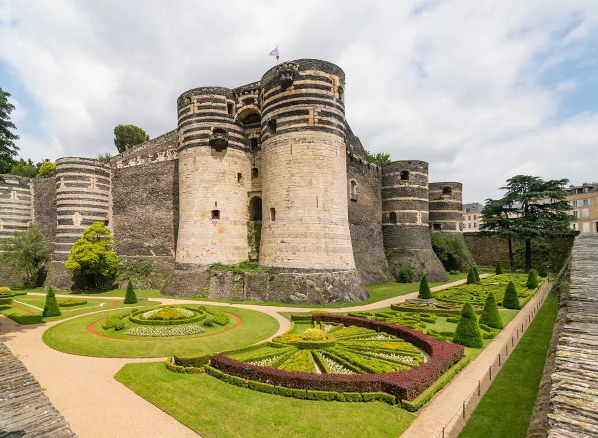 Ch&acirc;teau m&eacute;di&eacute;val avec des tours de pierre ray&eacute;es et des jardins manucur&eacute;s aux motifs g&eacute;om&eacute;triques sous un ciel nuageux.