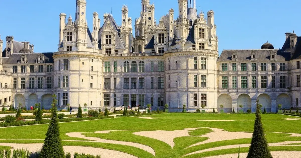 Un grand château avec des tours ornées et un jardin entretenu avec des pelouses et des allées géométriques.