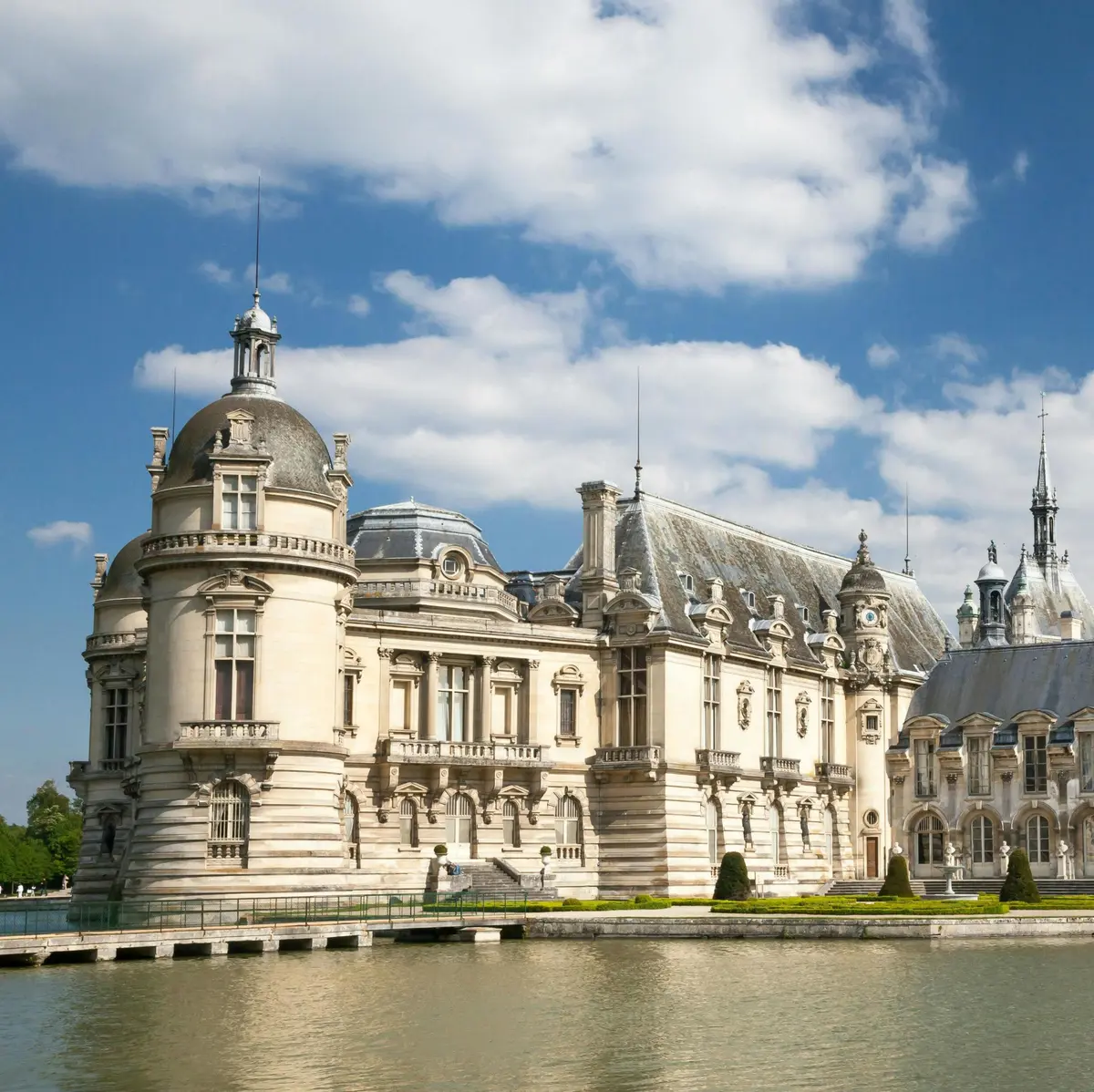 Un château historique orné de tourelles et de détails complexes se trouve au bord d'un plan d'eau calme sous un ciel partiellement nuageux.