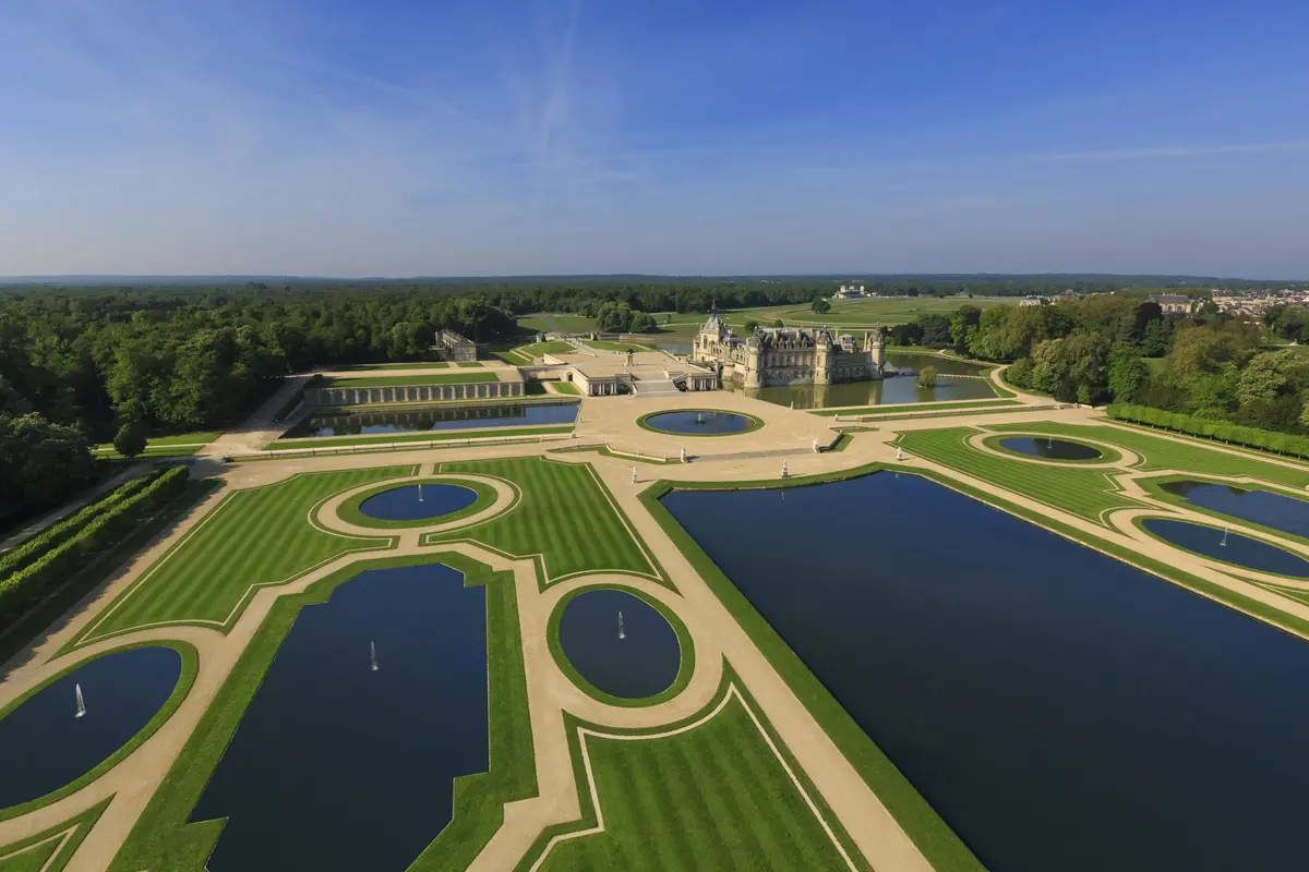 Vue aérienne d'un grand domaine avec des jardins à la française, des pièces d'eau géométriques et un château central entouré de vastes pelouses.