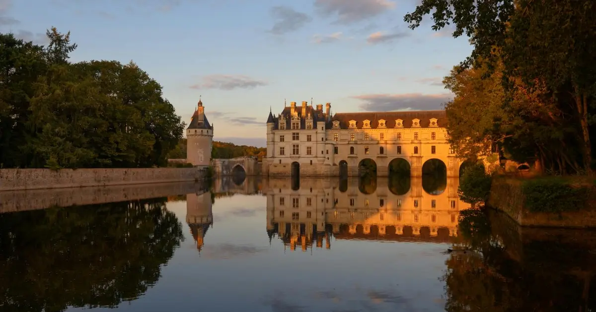 Château avec des ponts en arc au-dessus d'une rivière réfléchissante, des arbres de part et d'autre, et un ciel nuageux éclairé par un coucher de soleil.