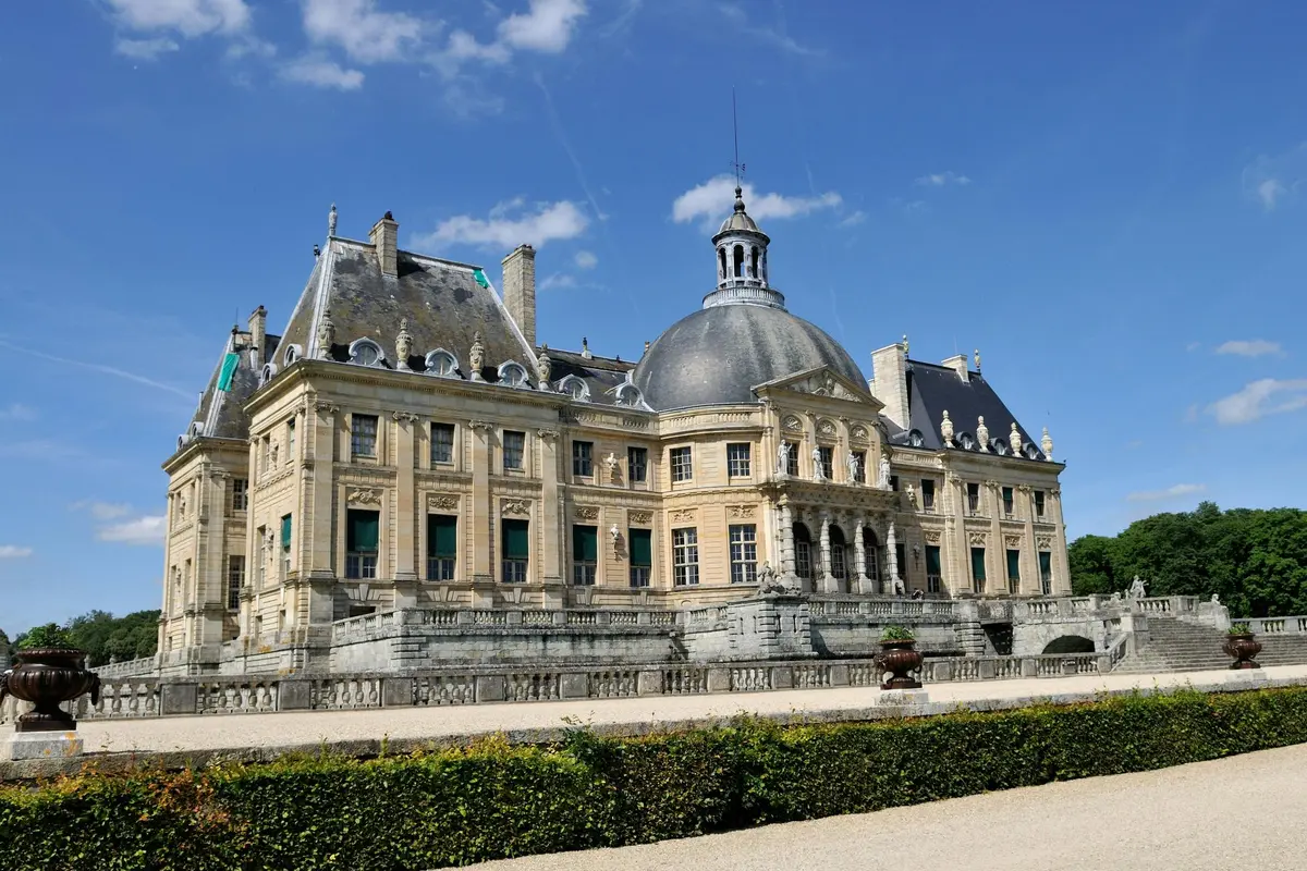 A grand historic chateau with a large dome, ornate architectural details, and manicured hedges in front under a clear blue sky.