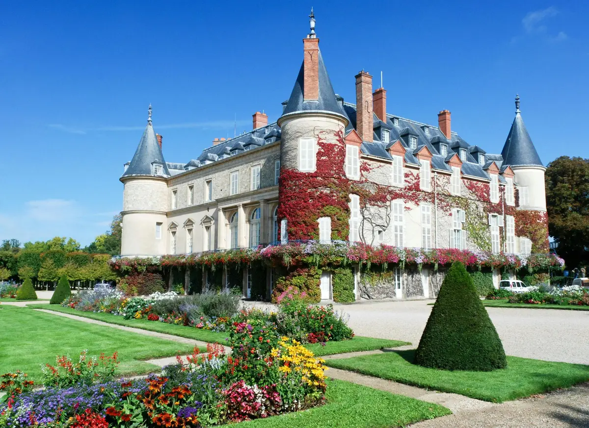 Un bâtiment historique dont les murs sont recouverts de lierre, entouré d'un jardin soigné avec des fleurs colorées et un arbuste taillé.