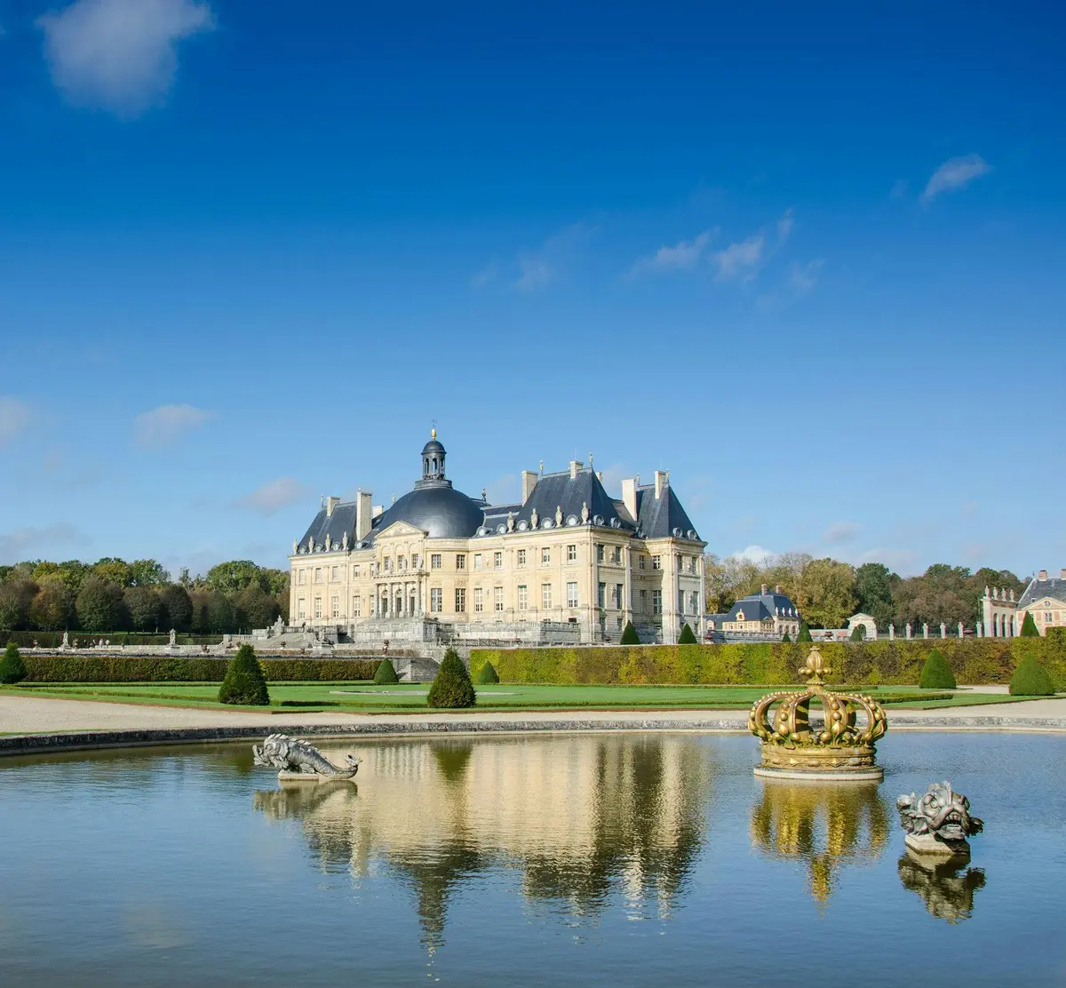 Château élégant avec jardins soignés et statues se reflétant dans un étang au premier plan sous un ciel bleu clair.