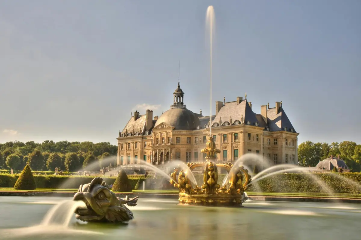 Un grand château avec des fontaines ornées au premier plan, avec des statues dorées et des jets d'eau sous un ciel clair.