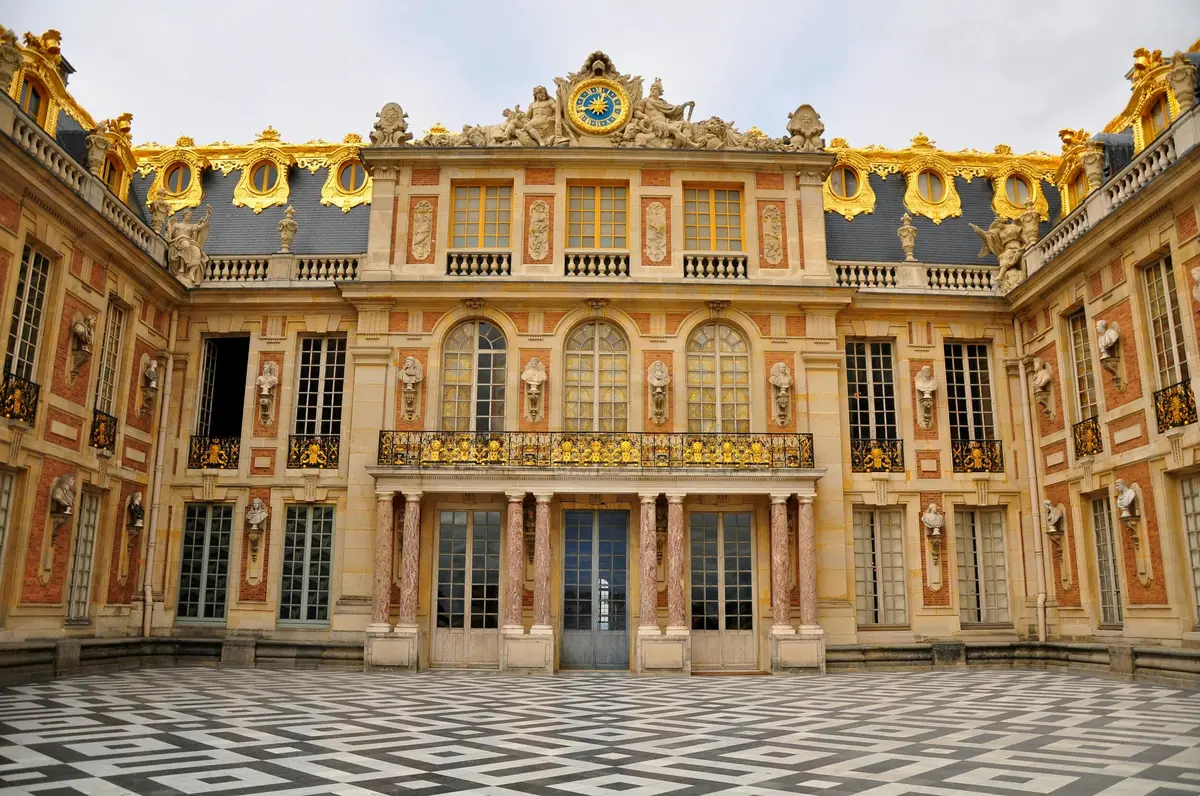 Ornate building facade with gold embellishments, arched windows, and checkered courtyard pattern, under a partly cloudy sky.