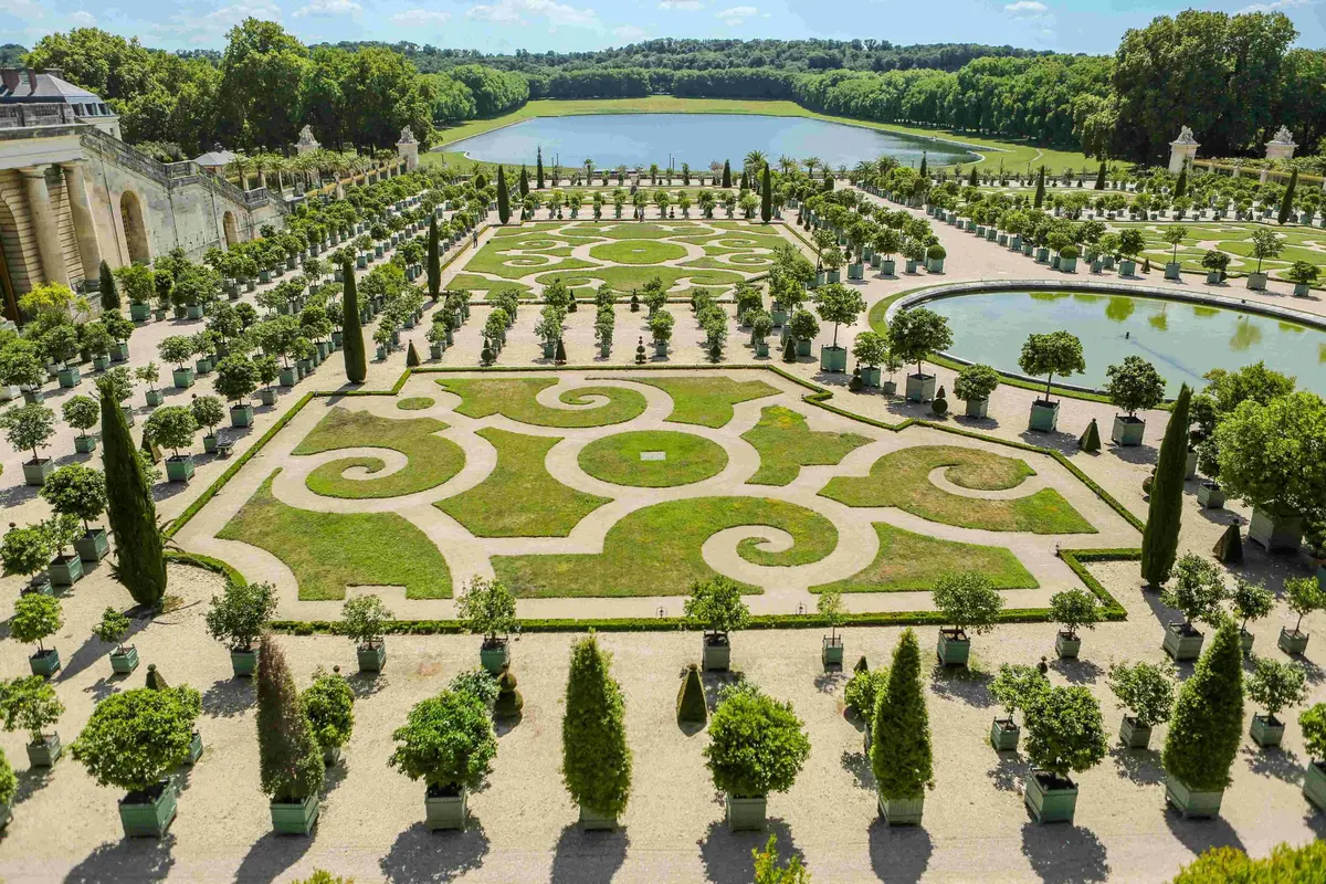 Jardin orné de haies géométriques symétriques, d'arbres en pot et d'un étang central, avec une forêt et un ciel en arrière-plan.